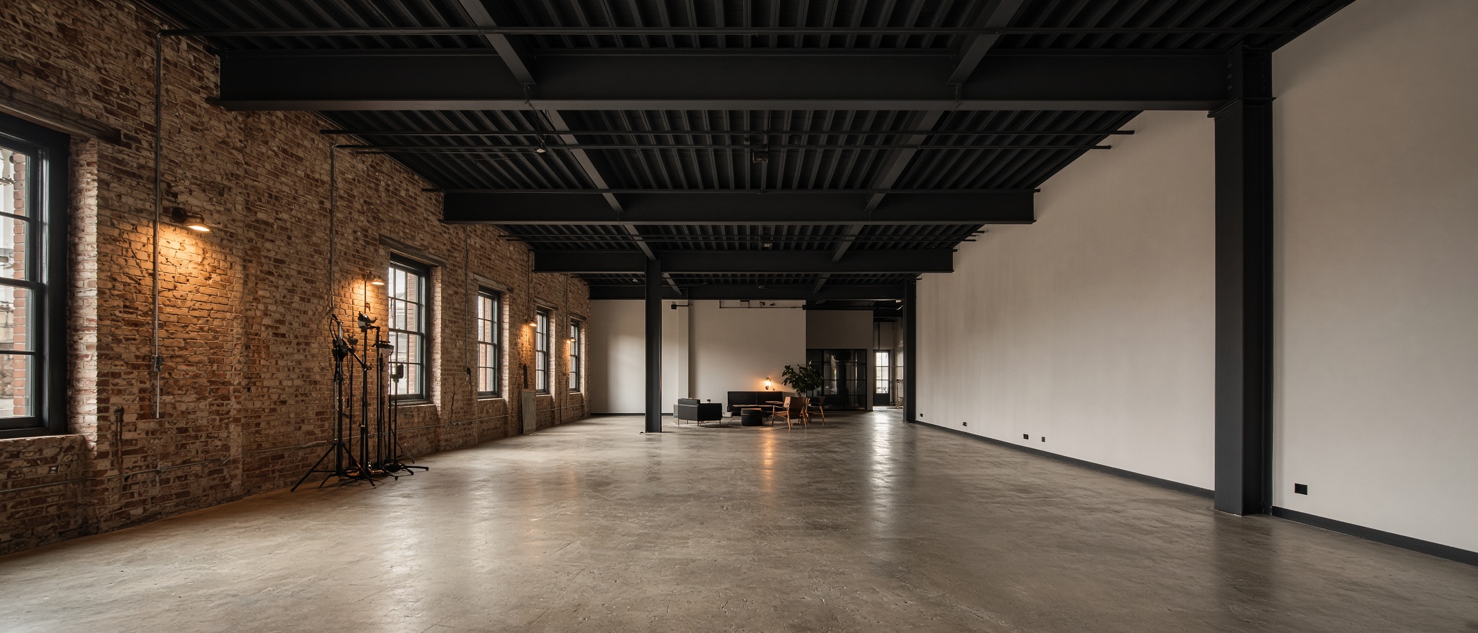 Wide view of the studio volume — high steel-beam ceiling, warm brick wall, bone plaster opposite, furniture set in the distance and open shooting floor.